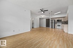 Open-concept living space featuring wood-finish flooring, a kitchen with stainless steel appliances, light wood-finish cabinetry, and a dark tile backsplash - 