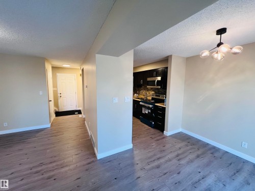 Entryway and living space featuring wood-finish flooring, a modern multi-bulb ceiling fixture, a white paneled entry door, and a kitchen with dark cabinetry, stainless steel appliances, and a tiled backsplash - 2544 79 Street, Edmonton, AB - Indoor
