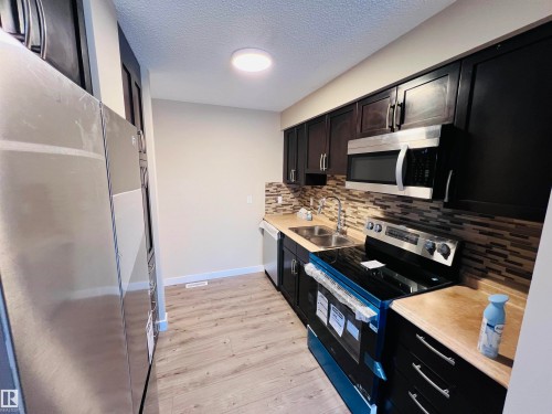 Kitchen featuring dark wood-finish cabinetry with silver hardware, stainless steel appliances, a multi-tone tile backsplash, light wood-finish countertops, and wood-finish flooring - 2544 79 Street, Edmonton, AB - Indoor Photo Showing Kitchen With Double Sink