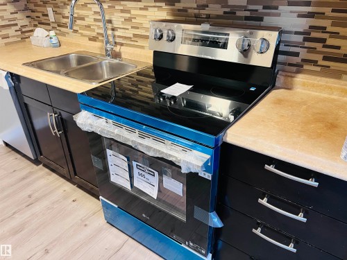 Kitchen with dark wood cabinetry, a mosaic tile backsplash, double basin stainless steel sink with a gooseneck faucet, light-toned countertops, and a new stainless steel and black range - 2544 79 Street, Edmonton, AB - Indoor Photo Showing Kitchen With Double Sink
