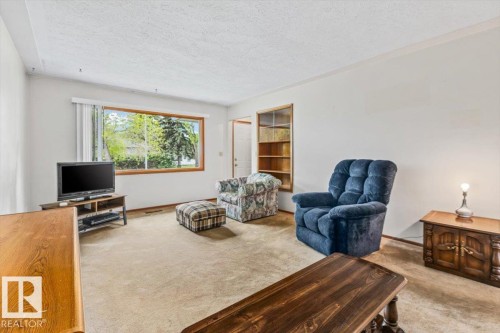Living area featuring a large picture window, light-colored carpeting, white walls, and a built-in wooden bookcase - 12923 113A Street, Edmonton, AB 