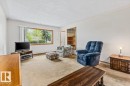 Living area featuring a large picture window, light-colored carpeting, white walls, and a built-in wooden bookcase - 12923 113A Street, Edmonton, AB 