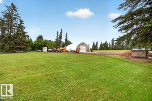 Expansive green lawn with mature evergreen trees, a large arched structure featuring a roll-up garage door, and a rustic wooden covered shelter - 572004 Highway 857, Rural Two Hills County, AB 