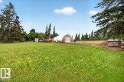 Expansive green lawn with mature evergreen trees, a large arched structure featuring a roll-up garage door, and a rustic wooden covered shelter - 