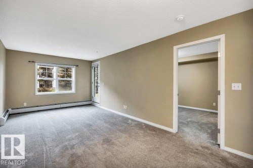 Spacious room featuring neutral-toned walls, gray carpeting, a large window, a glass-paneled door, and white trim - 116 630 Mcallister Loop, Edmonton, AB 