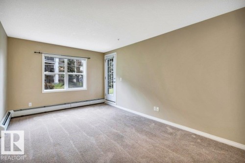 Carpeted room featuring a large window, a glass-paneled door, baseboard heating, and neutral-toned walls - 116 630 Mcallister Loop, Edmonton, AB 