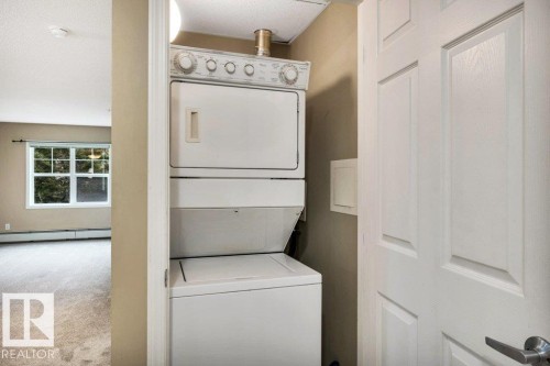White paneled door opening to a stacked white washer and dryer unit, featuring a light tan wall and off-white carpet flooring - 116 630 Mcallister Loop, Edmonton, AB 