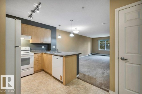 Functional kitchen featuring wood cabinetry, white appliances, a peninsula with sink, and track lighting, opening to a carpeted living space with a window - 116 630 Mcallister Loop, Edmonton, AB 