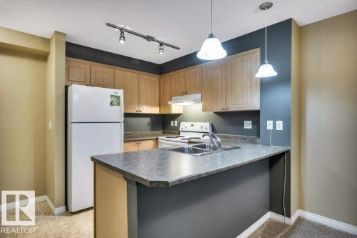 Kitchen featuring wood cabinetry, white appliances, a double basin sink, and a breakfast bar - 116 630 Mcallister Loop, Edmonton, AB 
