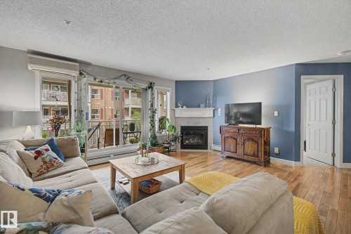Living area featuring wood-finish flooring, a corner fireplace with a white mantel, and extensive natural light from multiple windows - 308 10933 124 Street, Edmonton, AB 
