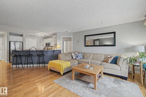 Open-concept living space featuring wood-finish flooring, a kitchen island with seating, stainless steel appliances, and white cabinetry - 308 10933 124 Street, Edmonton, AB 