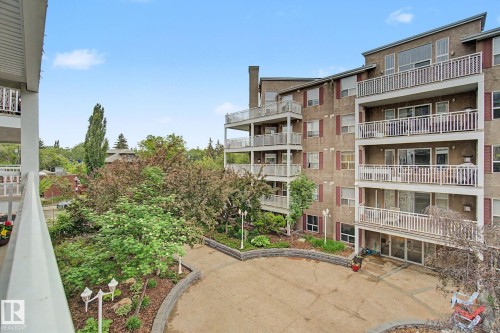 Multi-story residential building featuring balconies with white railings, a textured facade with red window shutters, and a paved courtyard with perimeter landscaping - 308 10933 124 Street, Edmonton, AB 