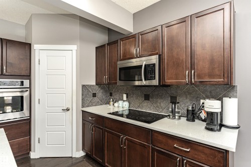 Kitchen featuring dark wood cabinetry, stainless steel appliances including a built-in microwave and oven, a white countertop, and a dark tile backsplash with accent squares - 115 57 Street, Edmonton, AB 