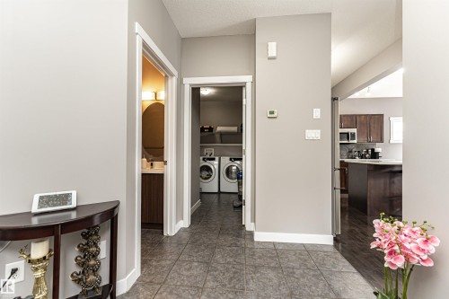 Tiled entryway flooring with access to a laundry area, a bathroom with an oval mirror, and a kitchen featuring dark wood cabinetry and a peninsula - 115 57 Street, Edmonton, AB 