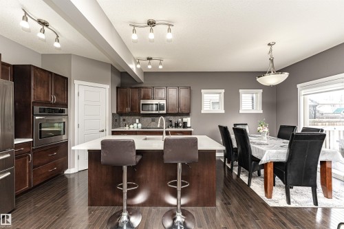 Open-concept kitchen and dining area featuring rich wood-finish cabinetry, stainless steel appliances, a center island with a white countertop, dark wood-finish flooring, and track lighting fixtures - 115 57 Street, Edmonton, AB 
