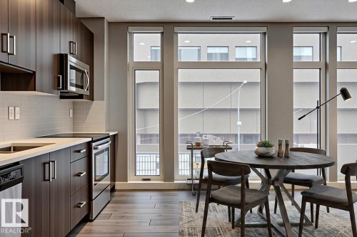 Modern kitchen and dining area featuring wood-finish flooring, dark wood-finish cabinetry, stainless steel appliances, and a light-toned tile backsplash - 401 10238 103 Street, Edmonton, AB - Indoor