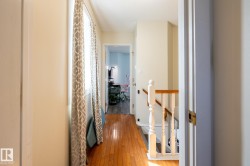 Hardwood flooring extends throughout this hallway, featuring a window with patterned drapes, and a staircase with a wood handrail and white balusters - 