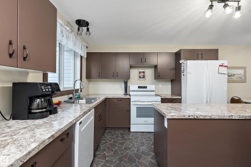 Kitchen featuring brown cabinetry, white appliances, a light-toned patterned countertop, a stainless steel sink with a gooseneck faucet, and flagstone-style flooring - 5210 55 Avenue, Wetaskiwin, AB 