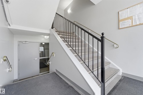 Stairwell featuring metal railings, wall-mounted handrails, and gray carpeted steps - #110 7805 159 St Nw, Edmonton, AB 