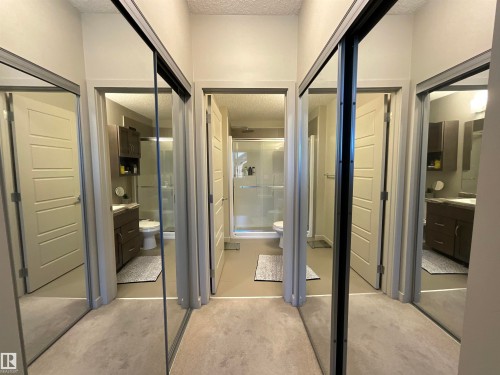 Mirrored sliding closet doors reflecting a modern bathroom with dark wood-finish cabinetry, a white rectangular sink, and a glass-enclosed shower - 209 1004 Rosenthal Boulevard, Edmonton, AB 