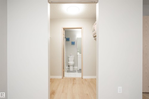 Hallway featuring light wood-finish flooring leading to a bathroom with a white toilet, white vanity, and wall-mounted mirror - 108 2508 50 Street, Edmonton, AB 