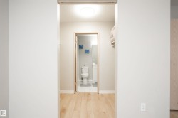 Hallway featuring light wood-finish flooring leading to a bathroom with a white toilet, white vanity, and wall-mounted mirror - 