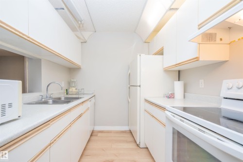 Galley kitchen featuring light wood-finish flooring, white cabinetry with subtle trim, white countertops, and white appliances including a refrigerator and range - 108 2508 50 Street, Edmonton, AB 