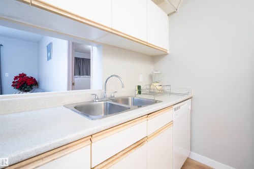 Kitchen featuring white cabinetry with wood-finish trim, a double basin stainless steel sink with a chrome faucet, light-colored countertops, and a built-in dishwasher - 108 2508 50 Street, Edmonton, AB 