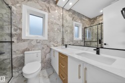 Bathroom featuring full-height marble-look wall tiling, a dual vanity with white cabinetry and wood-finish accents, integrated rectangular sinks, and matte black fixtures - 