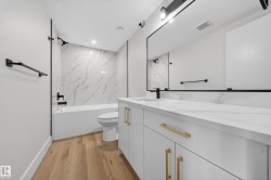 Bathroom featuring light wood-finish flooring, a white vanity with gold-tone hardware, a white countertop with integrated sink, a large framed mirror, and a tub with white and gray tiled surround - 