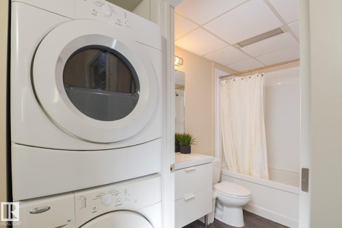 Dedicated laundry area featuring a stacked white washer and dryer, complemented by a full bathroom with a white vanity, integrated sink, and tub with shower - 411 10024 Jasper Avenue, Edmonton, AB 