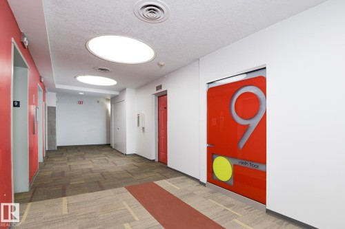 Brightly lit hallway featuring patterned carpet flooring, recessed circular lighting, red accent walls, and multiple elevator access points - 910 10024 Jasper Avenue, Edmonton, AB 
