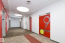 Brightly lit hallway featuring patterned carpet flooring, recessed circular lighting, red accent walls, and multiple elevator access points - 910 10024 Jasper Avenue, Edmonton, AB 