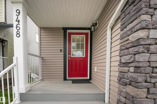 Welcoming red entry door with glass paneling, complemented by a covered porch with a light grey decking surface and white railings - 9468 209 St, Edmonton, AB 