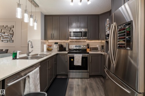 Modern kitchen featuring dark cabinetry, stainless steel appliances, light-toned countertops, a subway tile backsplash, and wood-finish flooring - 9468 209 St, Edmonton, AB 