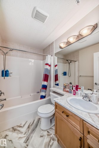 Bathroom featuring a white tub with shower, wood-finish vanity with a built-in sink, stone-look countertop, and a large mirror - 214 9938 104 Street, Edmonton, AB 