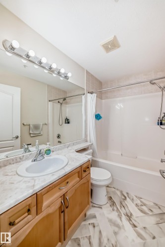Full bathroom featuring a single vanity with a white basin and light-colored countertop, wood-finish cabinetry, a large wall-mounted mirror, and a combined tub and shower unit with tile surround - 214 9938 104 Street, Edmonton, AB 