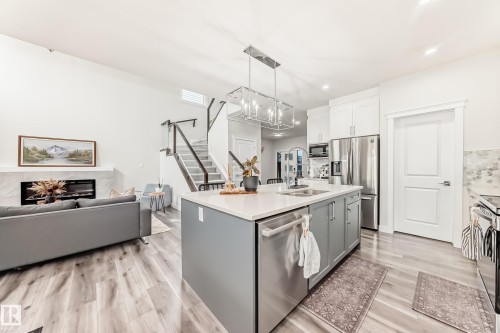 Spacious open-concept living area featuring a large kitchen island with a quartz countertop, stainless steel appliances, wood-finish flooring, a contemporary fireplace with a stone surround, and a modern staircase with glass panel railings - 589 Boulder Wynd, Leduc, AB - Indoor Photo Showing Kitchen With Double Sink With Upgraded Kitchen