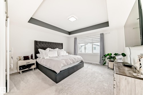 Spacious bedroom featuring plush carpeting, a large window with horizontal blinds and drapery, a tray ceiling with a dark accent, and a built-in ceiling light fixture - 589 Boulder Wynd, Leduc, AB - Indoor Photo Showing Bedroom