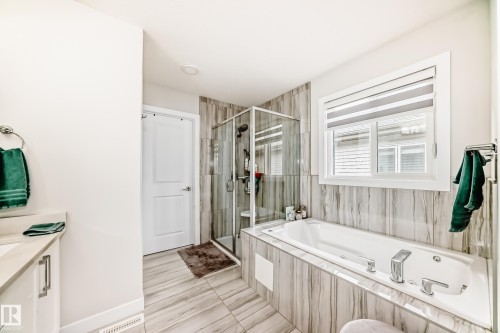 Bathroom featuring a large soaker tub, glass-enclosed shower, wall-mounted vanity with integrated sink, wood-finish tile flooring, and a large window with privacy blinds - 589 Boulder Wynd, Leduc, AB - Indoor Photo Showing Bathroom
