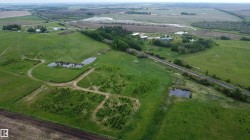 Expansive green landscape featuring multiple ponds, a residential structure with green roofing, a treeline, and a railroad track - 