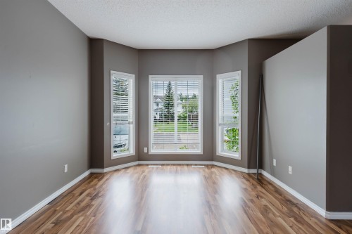 Bay window feature with three vertical windows and white blinds, complemented by wood-finish flooring, neutral gray walls, and white baseboards - 38 1780 Glastonbury Boulevard, Edmonton, AB 