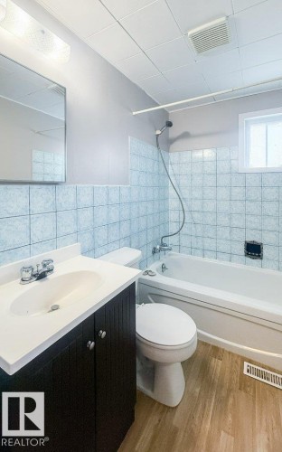 Full bathroom featuring a white bathtub with blue tile surround, a dark wood-finish vanity with an integrated white sink, and wood-finish flooring - 9836 100 Street, Westlock, AB 