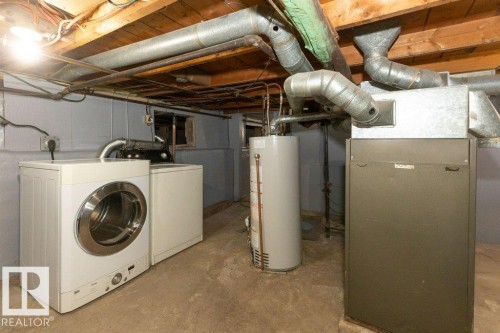 Utility area featuring exposed wood joists, gray painted walls, a concrete floor, a water heater, and a furnace with exposed ductwork - 9836 100 Street, Westlock, AB 