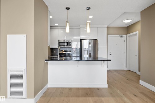 Contemporary kitchen featuring high-gloss white cabinetry, stainless steel appliances, dark countertop, and pendant lighting over a breakfast bar - 307 11080 Ellerslie Rd Sw, Edmonton, AB 