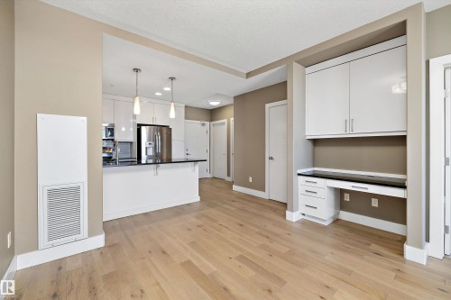 Light wood-finish flooring extending into an open-concept kitchen featuring white cabinetry, a dark countertop, and stainless steel appliances - 307 11080 Ellerslie Rd Sw, Edmonton, AB 