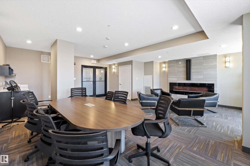Multifunctional office common area featuring a wood-finish conference table, contemporary linear fireplace with a textured surround, recessed lighting, wall sconces, and patterned carpet flooring. - 307 11080 Ellerslie Rd Sw, Edmonton, AB 