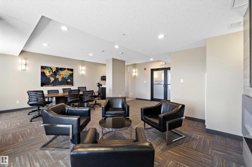 Multifunctional office common area featuring a wood-finish conference table, contemporary linear fireplace with a textured surround, recessed lighting, wall sconces, and patterned carpet flooring. - 307 11080 Ellerslie Rd Sw, Edmonton, AB 