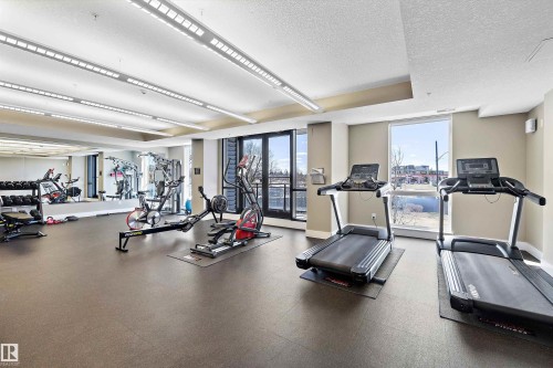 Fitness center featuring rubberized flooring, mirrored wall, multiple cardio machines, and strength training equipment. - 307 11080 Ellerslie Rd Sw, Edmonton, AB 
