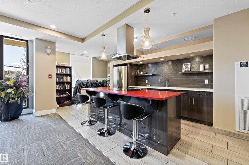 The Recreation Room has a modern kitchen featuring a striking red island countertop, stainless steel appliances, dark wood-finish cabinetry, and a dark subway tile backsplash, with a pool table and a lounge type seating to enjoy with your company. - 307 11080 Ellerslie Rd Sw, Edmonton, AB 